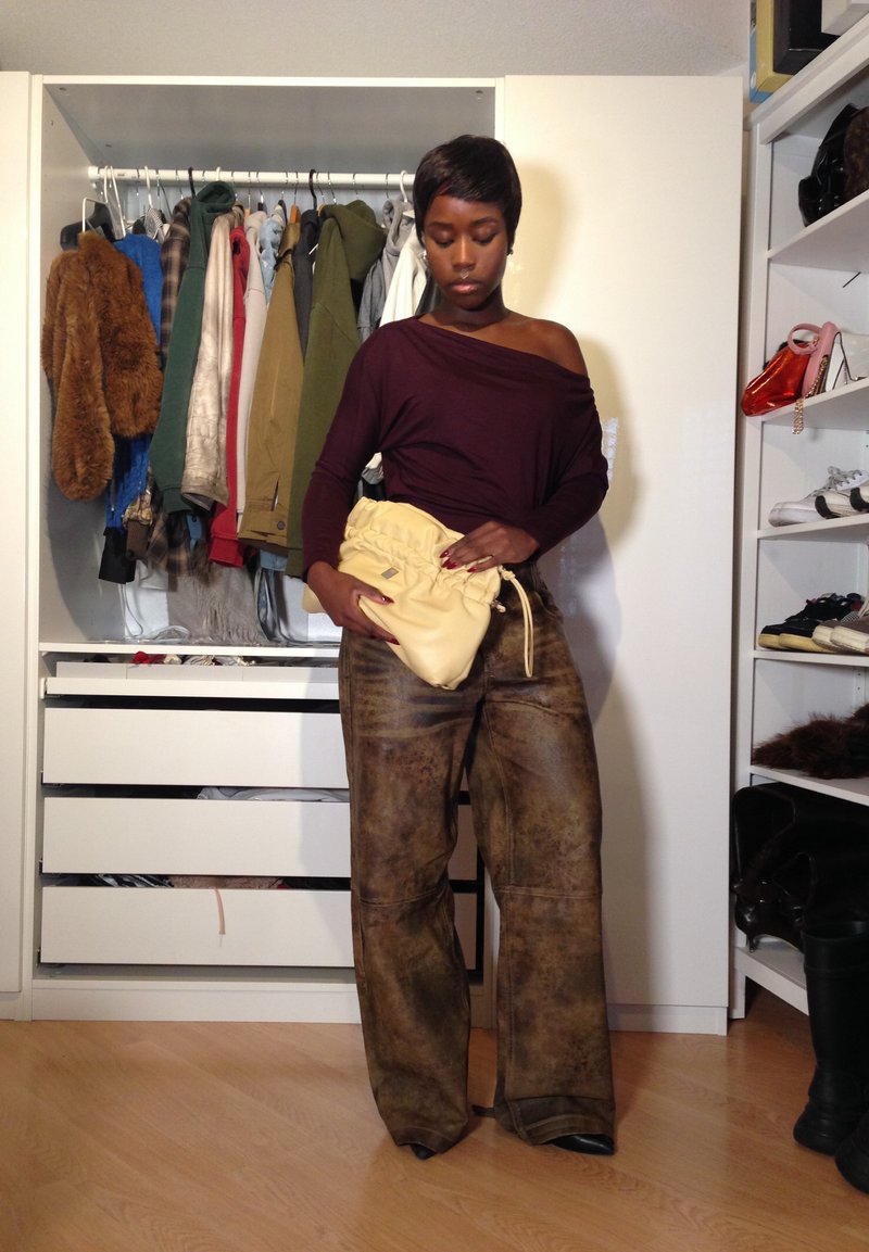 Donna con pantaloni larghi marroni e top bordeaux a spalle scoperte tiene in mano una borsa gialla arricciata, in piedi in una stanza con armadio aperto e ripiani.