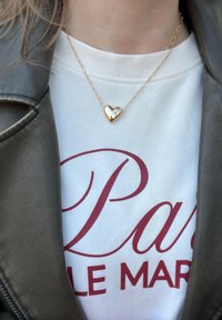 Un collier avec un pendentif en forme de cœur en or suspendu à une fine chaîne, porté sur un sweat-shirt blanc avec un texte bordeaux, sous une veste en cuir noir.