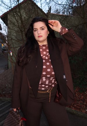 Mujer con cabello largo y oscuro, vestida con blusa marrón de lunares, blazer oscuro y pantalones, sosteniendo un bolso de cuadros, de pie al aire libre cerca de árboles sin hojas.