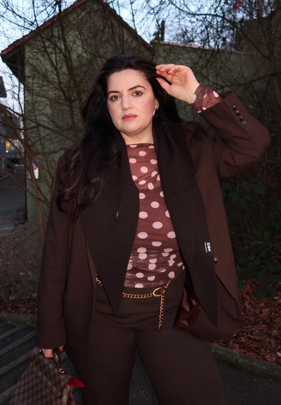 Mujer con cabello largo y oscuro, vestida con blusa marrón de lunares, blazer oscuro y pantalones, sosteniendo un bolso de cuadros, de pie al aire libre cerca de árboles sin hojas.