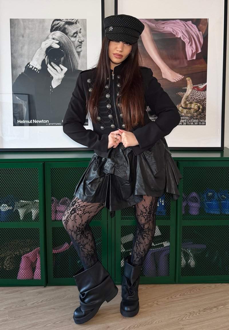 Vrouw in zwarte militair stijl jas, leren rok, panty's met patroon en laarzen die poseren binnen voor ingelijste kunst en groene schoenenkasten.