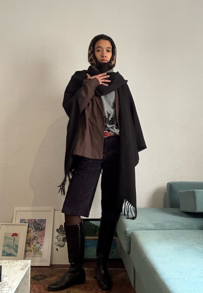 Personne debout à l'intérieur portant une veste marron, une écharpe noire, une capuche à imprimé léopard, un short jusqu'aux genoux et des bottes noires à côté d'un canapé bleu sarcelle et d'une œuvre encadrée.