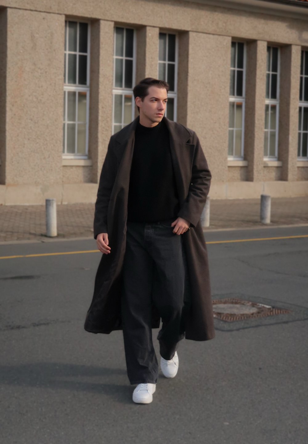 Casaco longo escuro sobre camisola preta e calças largas pretas. Ténis brancos. Ambiente urbano com parede de pedra e janelas ao fundo.