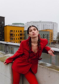 Rdeča lanena blazer in usklajene oprijete legice. Model pozira na balkonu z zgradbami v ozadju. Dolgi pleteni lasje in minimalni dodatki.