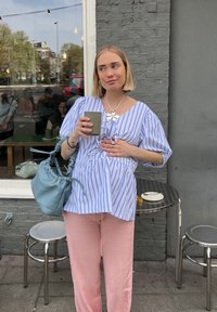 Ριγέ μπλουζάκι μπλε και λευκό, ροζ παντελόνια, κρατώντας ένα πράσινο φλιτζάνι και ένα διάφανο ποτήρι. Περιέχει μια ανοιχτό μπλε τσάντα; καθισμένα τραπέζι και καρέκλες στο υπόβαθρο.