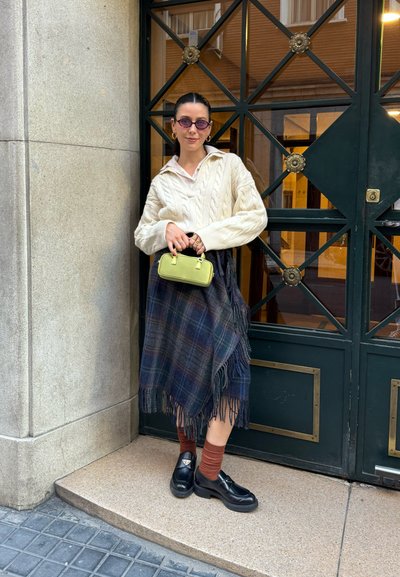 Suéter de punto trenzado crema, falda de cuadros con flecos en tonos oscuros, bolso verde con herrajes dorados, zapatos negros y calcetines marrones.