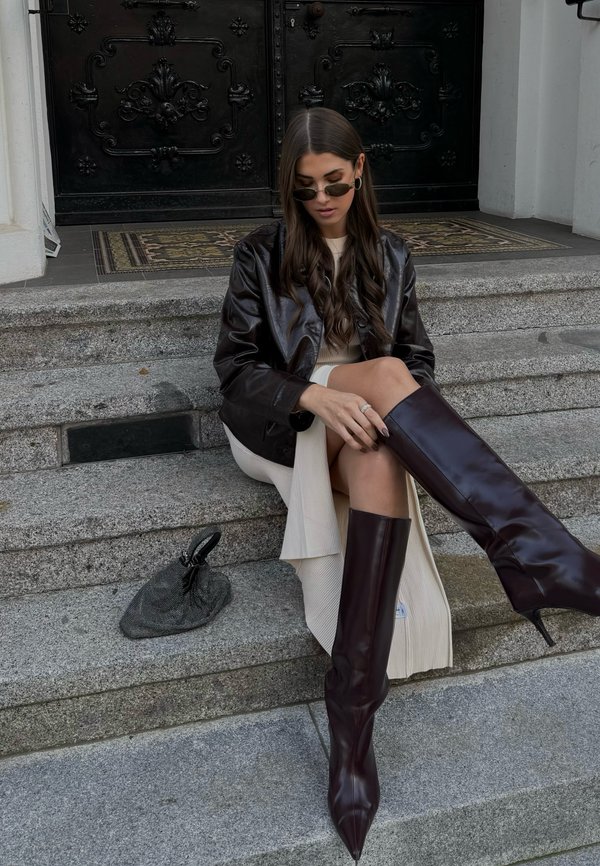 Bruine leren cropped jack, geribbelde crèmekleurige jurk, hoge bordeauxrode stiletto laarzen en een gestructureerde grijze handtas op stenen trappen met een zwarte deur.