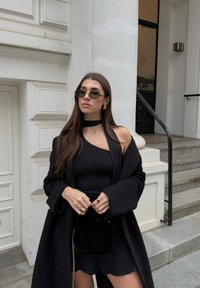 Černý oversized kabát s širokými klopami, přiléhavý černý top a volánková černá mini sukně. Doplňky zahrnují malou černou kabelku a sluneční brýle.