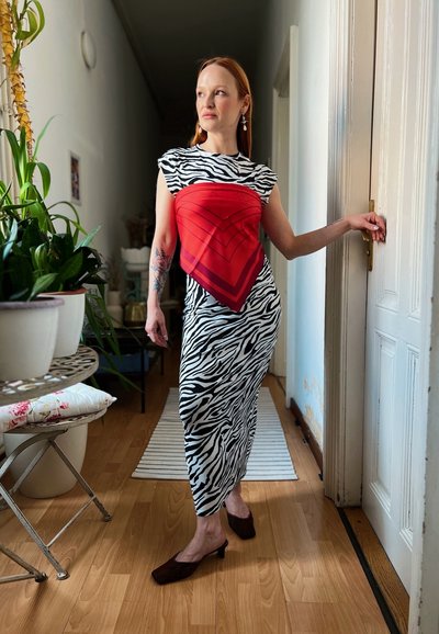 El vestido presenta un estampado de cebra en blanco y negro, con un acento triangular en rojo. La modelo lleva unos zapatos de tacón marrones y está de pie en un entorno indoor.