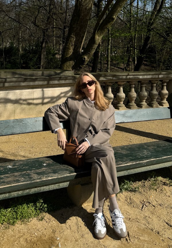 Beige Blazer mit großen Knöpfen über einem passenden Rock. Braune Handtasche, weiße Sneakers mit grauen Akzenten, die auf einer grünen Bank sitzen.