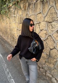 Fekete kötött pulóver, szürke bő szárú farmer, fekete kézitáska ezüst szegecsekkel, napszemüveg, egy texturált kőfal előtt.