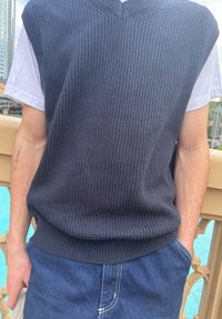 Suéter sin mangas de punto en un oscuro tono azul marino con escote en V y textura acanalada, llevado sobre una camiseta blanca, combinado con pantalones de mezclilla azul.