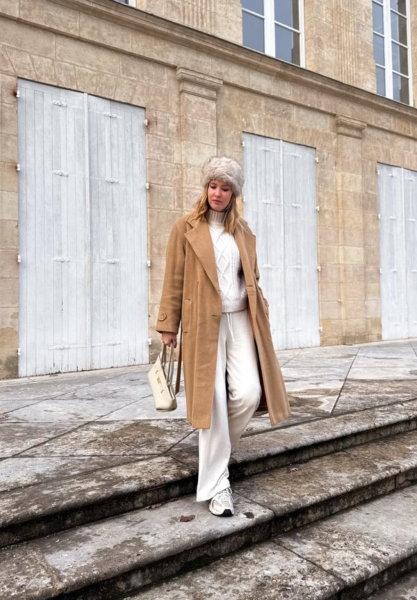 Sieviete nolaižas pa akmens kāpnēm ārpus bēša krāsas ēkas ar aizvērtām žalūzijām, valkājot brūnganu mēteli, baltas bikses, adītu džemperi, pūkainu cepuri un sporta apavus.