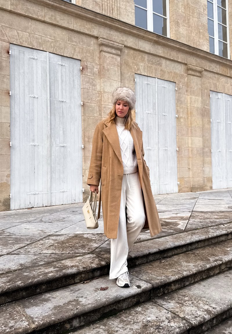 Γυναίκα κατεβαίνει πέτρινα σκαλοπάτια έξω από μπεζ κτίριο με κλειστά παντζούρια, φορώντας καμηλό παλτό, άσπρο παντελόνι, πλεκτό πουλόβερ, γούνινο καπέλο και αθλητικά παπούτσια.