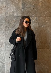 Zwart dubbelgeknoopt mantel, grijze top eronder, zwarte crossbodytas met gestructureerde accenten, donkere zonnebril, tegen een achtergrond van marmeren muur.