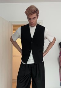 Jeune personne aux cheveux roux clair portant un gilet noir sur une chemise blanche et un pantalon large noir, debout à l'intérieur les mains sur les hanches.
