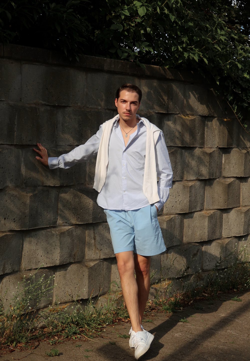 Calções azul claro, camisa branca com riscas, toalha branca pousada sobre os ombros. Apenado contra uma parede de pedra texturizada. Folhagem verde acima.