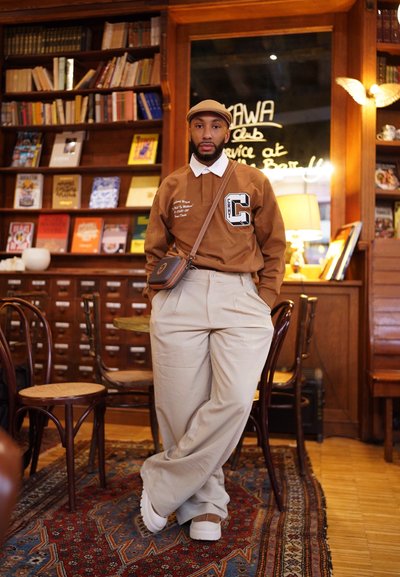 Camisa marrón de manga larga con un cuello blanco, pantalones de tela crema con pliegues y zapatillas blancas. Presenta un bolso cruzado y un fondo de paneles de madera.