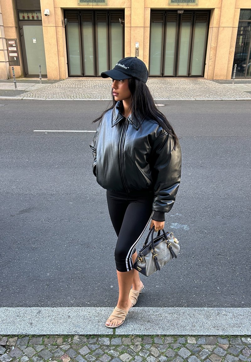 Giacca bomber in pelle nera, leggings neri aderenti con strisce bianche, sandali beige e una borsa grigia. In piedi su un marciapiede.