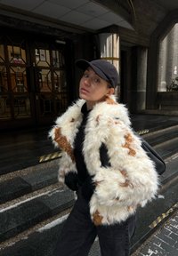 Jeune femme portant une casquette noire et une veste en fourrure blanche et brune, debout à l'extérieur près des marches d'un bâtiment avec des portes sombres et des colonnes réfléchissantes.