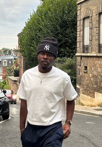 Λευκό κοντομάνικο πουκάμισο, μαύρο σκουφάκι με λευκό λογότυπο, χρυσό περιδέραιο, ναυτική μπλε παντελόνα, περπατώντας σε έναν δρόμο με πρασινάδα και κτήρια.