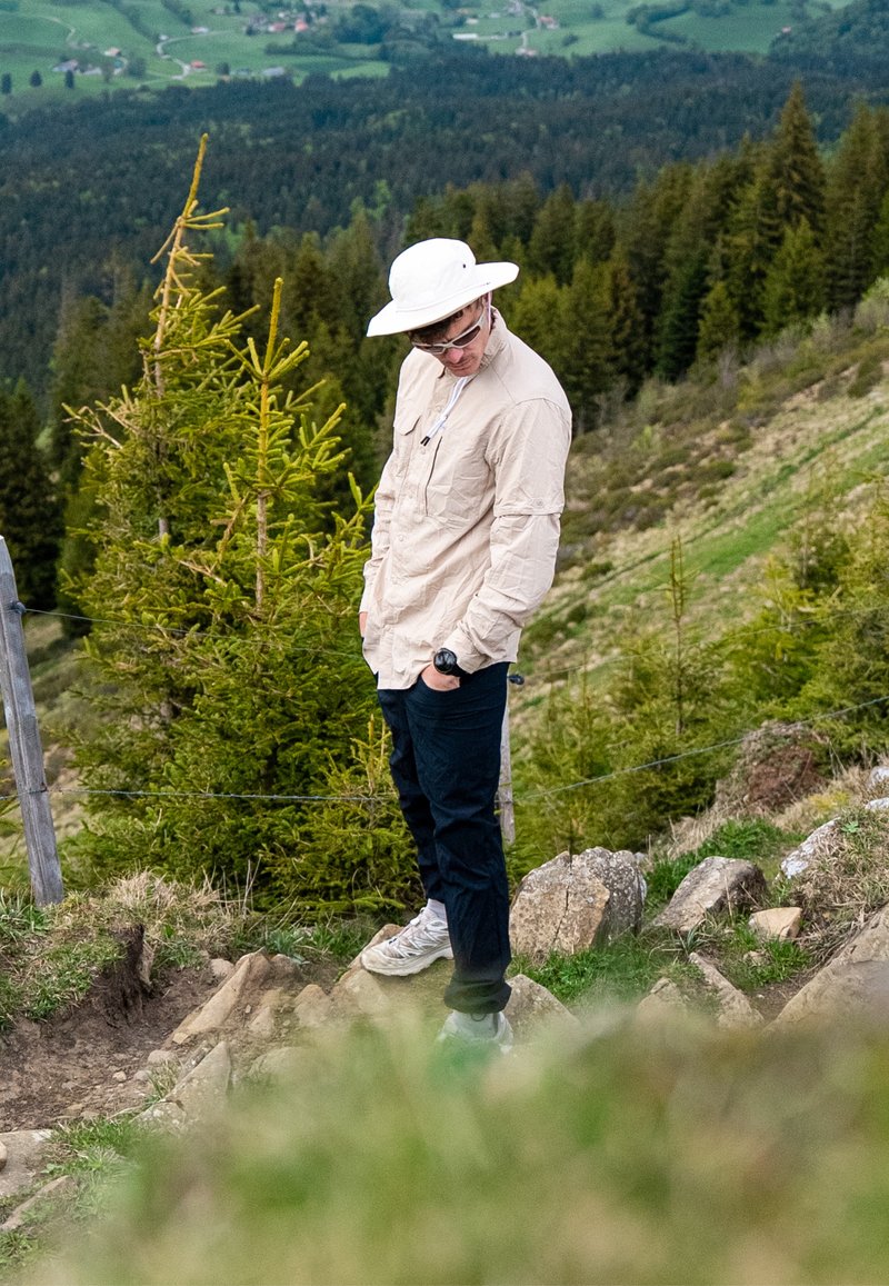 Personne en chemise claire, chapeau de soleil blanc et lunettes de soleil, debout sur un chemin rocailleux sur la colline, entourée d’arbres à feuilles persistantes avec des champs au loin en contrebas.