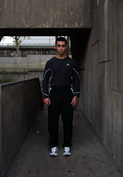 Un joven vestido con ropa deportiva negra y zapatillas blancas está de pie en un estrecho pasillo de concreto bajo un paso elevado.