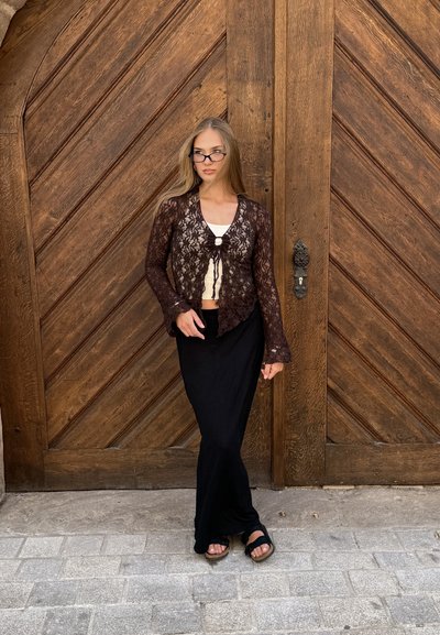 Cardigan de encaje en marrón oscuro con mangas acampanadas y cierre con lazo sobre una blusa clara, combinado con una falda negra y sandalias, frente a una puerta de madera.