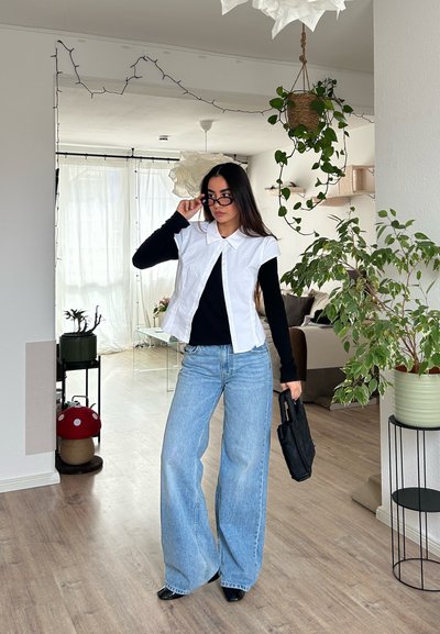 Mujer ajustando gafas negras, lleva una camisa blanca sin mangas sobre un top negro, vaqueros anchos azules, sosteniendo un bolso negro en una sala luminosa con plantas.