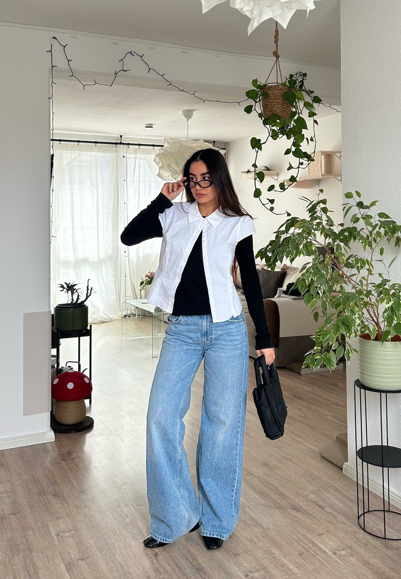 Femme ajustant des lunettes noires, vêtue d'une chemise blanche sans manches sur un top noir, jean bleu large, tenant un sac noir dans un salon lumineux avec des plantes.