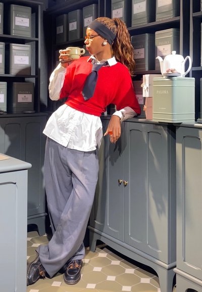 Suéter de punto rojo sobre una camisa blanca, corbata negra, pantalones de pierna ancha grises y zapatos marrones. Sosteniendo una taza, de pie contra tinajas de almacenamiento verdes.