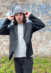 Černý blazer s texturovanou tkaninou, šedé tričko a černé tepláky. Šedý pletený beanie na hlavě. Stěna s modrým a bílým textem v pozadí.