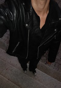 Zwarte leren jas met ritsen en zilveren sierbeslag, gedragen over een zwart shirt. Zwarte broek en schoenen met witte accenten zichtbaar.