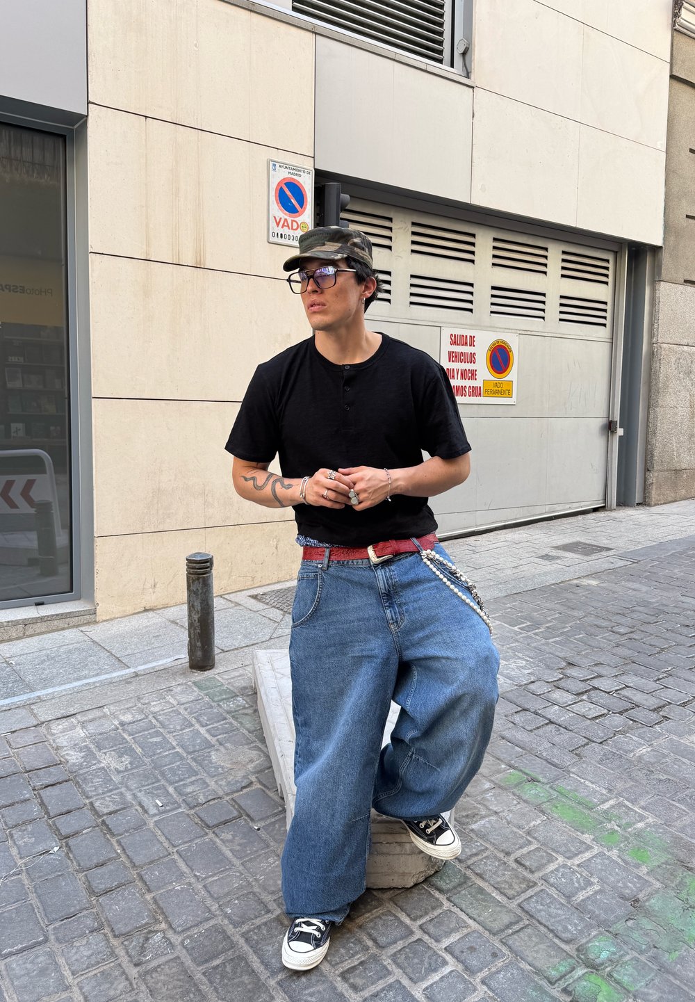 Fekete rövid ujjú henley póló, oversized kék farmernadrág, piros öv és fekete sportcipők. Kiegészítők közé tartoznak láncok és szemüveg. Szabadtéri környezet.