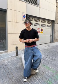 Fekete rövid ujjú henley póló, oversized kék farmernadrág, piros öv és fekete sportcipők. Kiegészítők közé tartoznak láncok és szemüveg. Szabadtéri környezet.