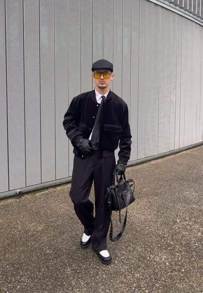 Chaqueta bomber de terciopelo negro con una camisa y corbata blanca, pantalones negros, guantes de piel, gafas de sol amarillas y un bolso negro. Textura suave.