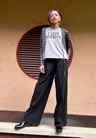 Camiseta gris con el texto "ME ENCANTAN LOS COWBOYS", superpuesta a una camisa de manga larga larga, a rayas y transparente. Pantalones anchos de rayas negras y zapatos negros.