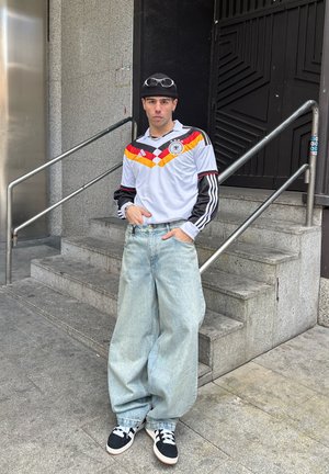 Joven con gorro negro, camiseta retro de fútbol de Alemania, jeans anchos de color azul claro y zapatillas negras, de pie junto a unas escaleras de concreto.