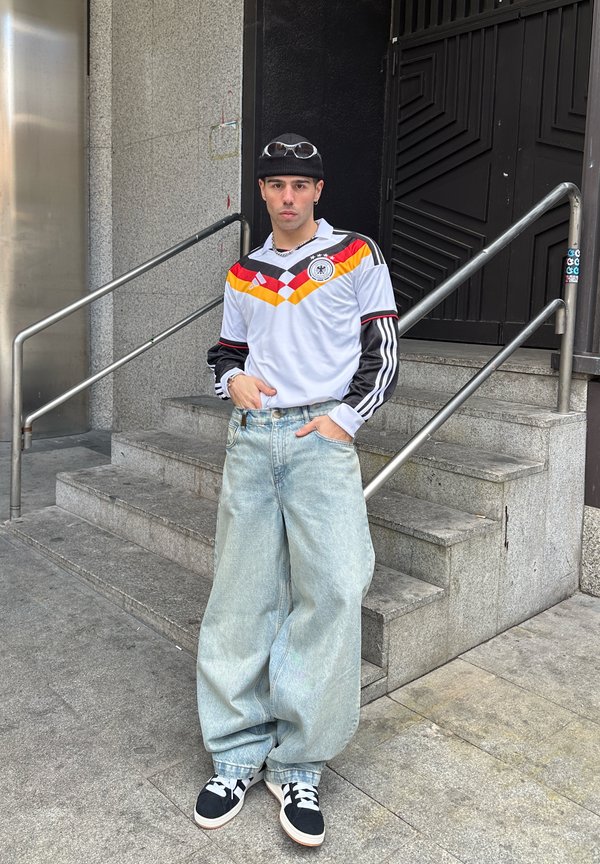 Jonge man met een zwarte beanie, een retro Duits voetbalshirt, wijde lichtblauwe spijkerbroeken en zwarte sneakers staand bij betonnen trappen.