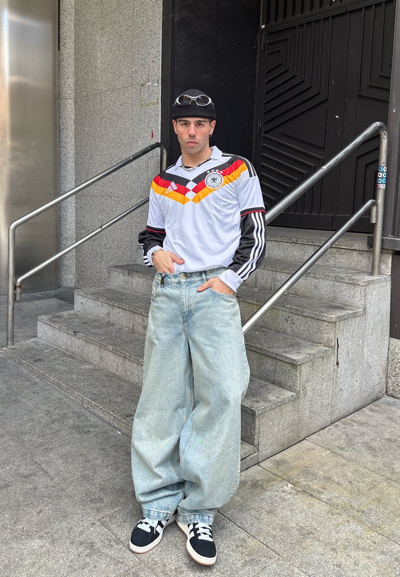 Jeune homme portant un bonnet noir, un maillot de football rétro de l'Allemagne, un jean large bleu clair et des baskets noires, debout près d'un escalier en béton.