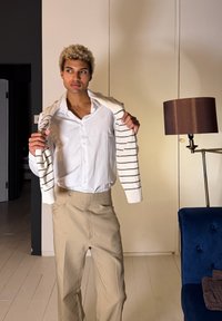 Νεαρός με σγουρά ξανθά μαλλιά προσαρμόζει την ριγέ μπλούζα πάνω από λευκό πουκάμισο και μπεζ παντελόνι σε ένα μοντέρνο δωμάτιο με λάμπα και μπλε καρέκλα.