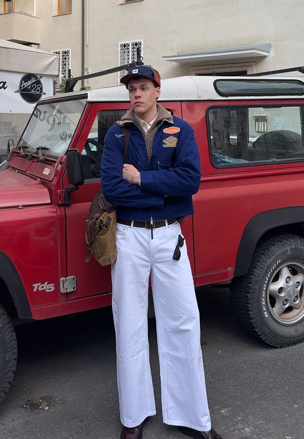 Young man in blue jacket, white wide-leg pants, and cap stands with arms crossed in front of a red Land Rover Defender Td5 vehicle.
