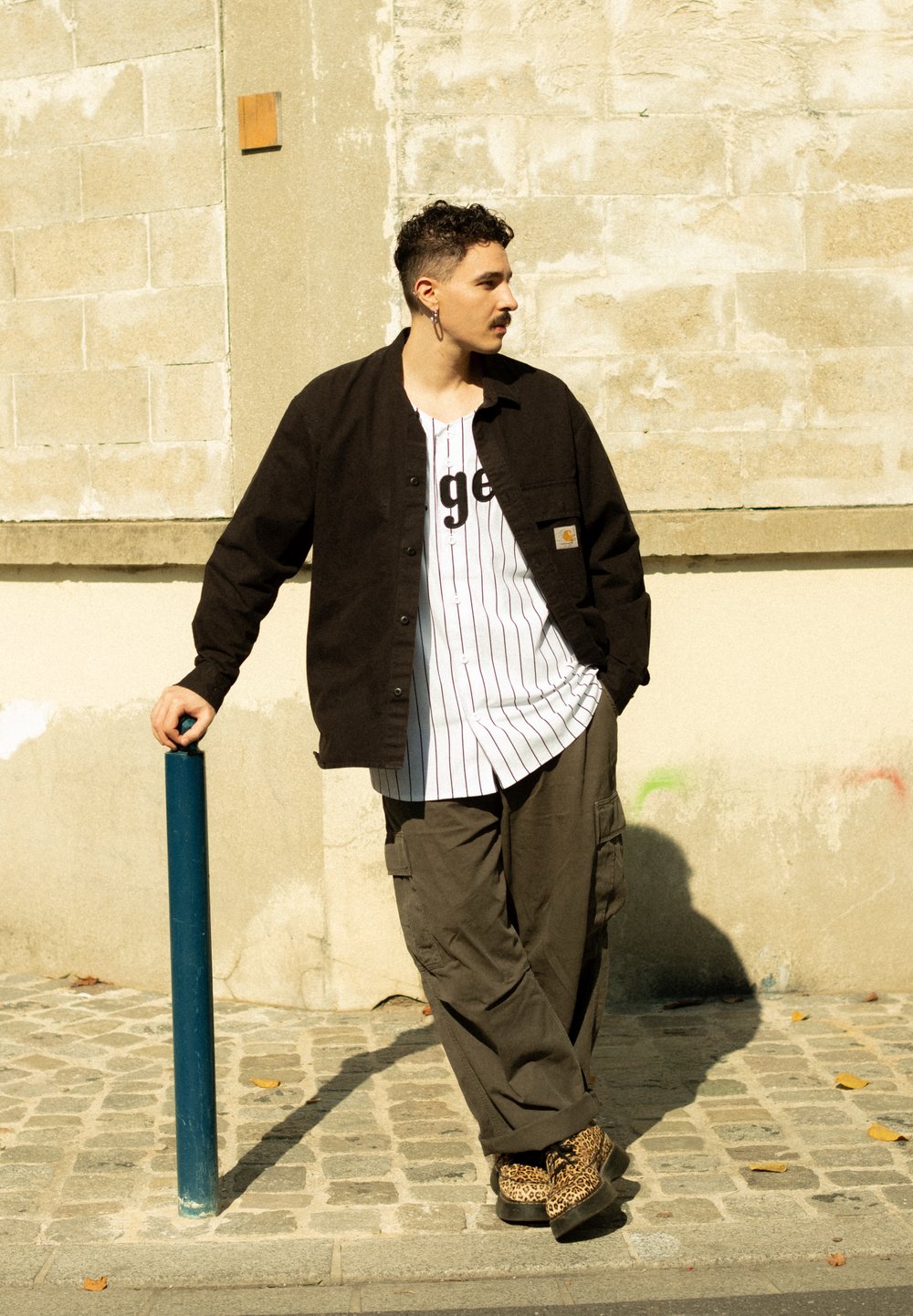 Nero camicia con bottoni, t-shirt bianca a righe sottostante con scritte nere, pantaloni cargo verde oliva e scarpe marroni con stampa leopardata. Polo blu nelle vicinanze.