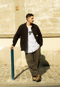 Nero camicia con bottoni, t-shirt bianca a righe sottostante con scritte nere, pantaloni cargo verde oliva e scarpe marroni con stampa leopardata. Polo blu nelle vicinanze.
