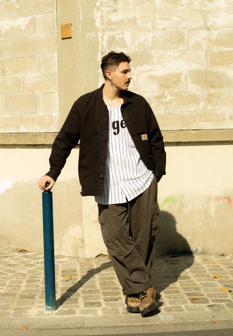 Schwarzes Hemd mit Knöpfen, darunter ein weißes T-Shirt mit Streifen und schwarzer Schrift, olivfarbene Cargo-Hosen und braune Schuhe mit Leopardenmuster. Nahebei ein blauer Pfosten.