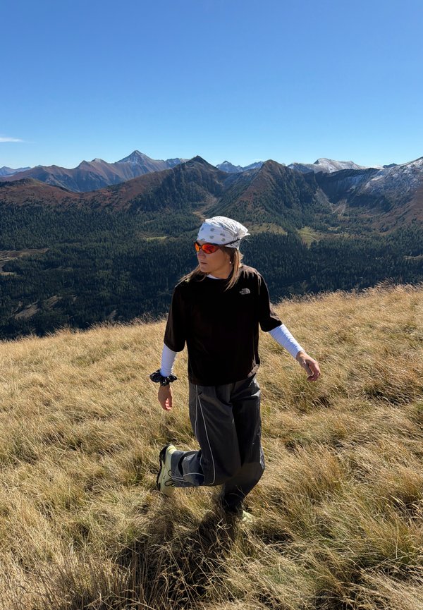 Un randonneur portant un bandana et des lunettes de soleil marche dans une prairie alpine herbeuse, avec des montagnes boisées sous un ciel bleu dégagé.