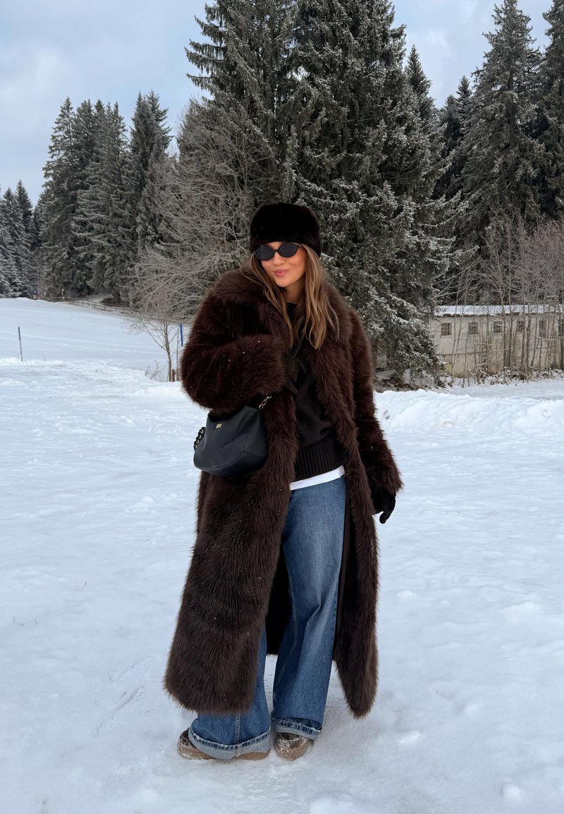 Femme portant un manteau en fourrure brun foncé, un chapeau, des lunettes de soleil et un jean bleu, debout sur un sol enneigé avec des sapins et un bâtiment en arrière-plan.