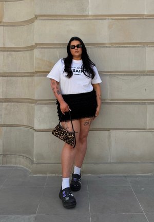 Camiseta gráfica blanca, mini falda negra con volantes, bolso de mano con estampado de leopardo, zuecos negros decorados con calcetines blancos, de pie contra una pared texturizada.