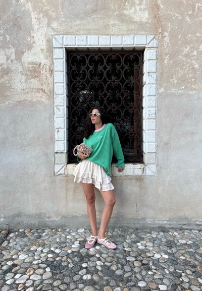 Sudadera de gran tamaño a rayas verdes con ribete rosa, falda blanca con volantes en capas, y zapatos rosas con tiras blancas, de pie junto a una ventana decorativa de hierro.