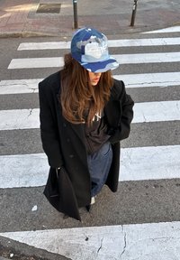 Μαύρο διπλό κουμπωτό παλτό, καμουφλάζ καπέλο με λογότυπο, μαύρη φούτερ μπλούζα, μπλε τζιν παντελόνι, μαύρες ψηλές μπότες, στέκεται σε διάβαση.